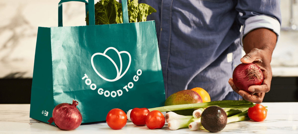 Borsa Too Good To Go con verdure fresche e mano che tiene una cipolla rossa.