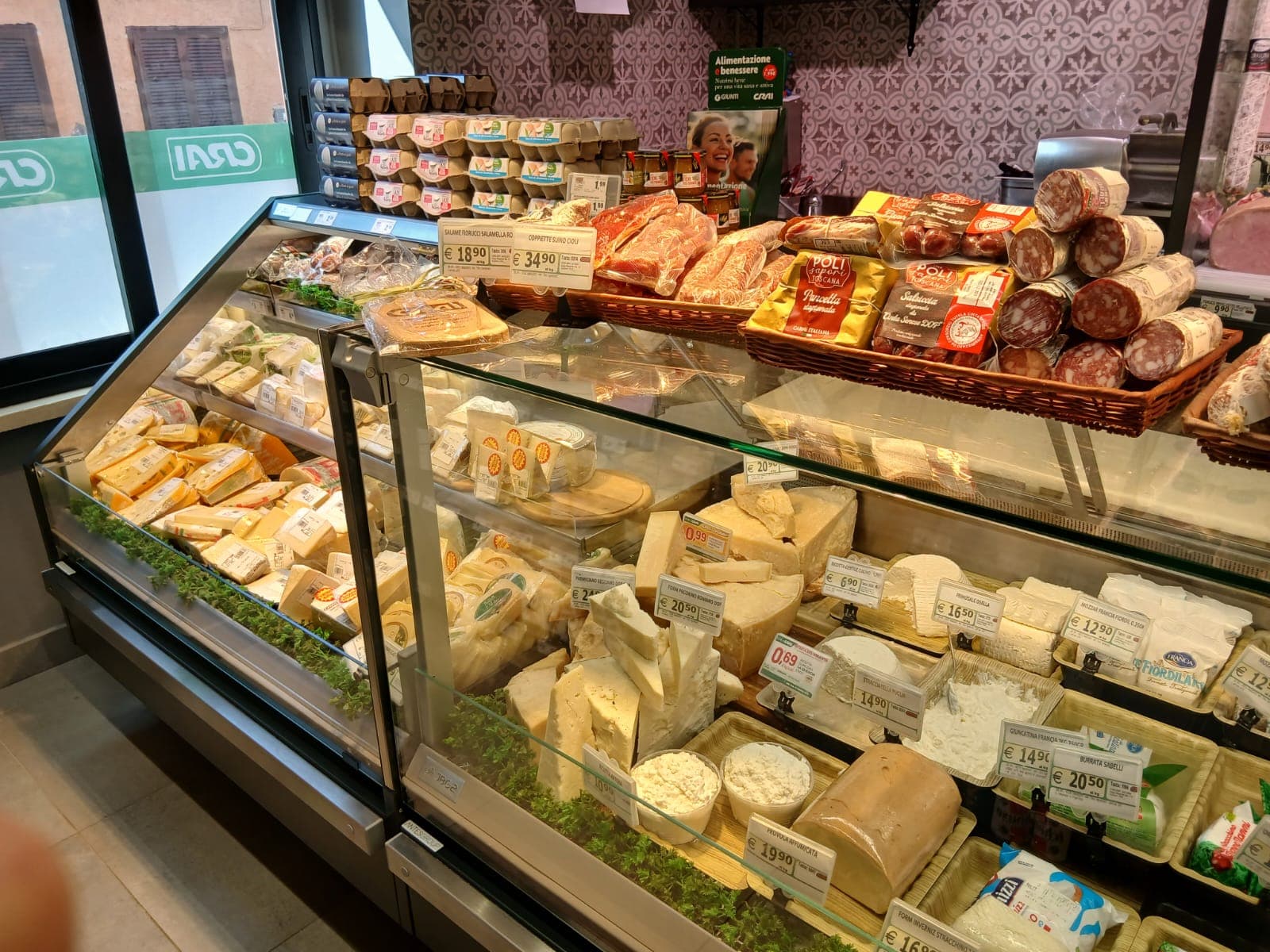 Vetrina refrigerata con vari formaggi, salumi tipici e confezioni di uova in un supermercato.