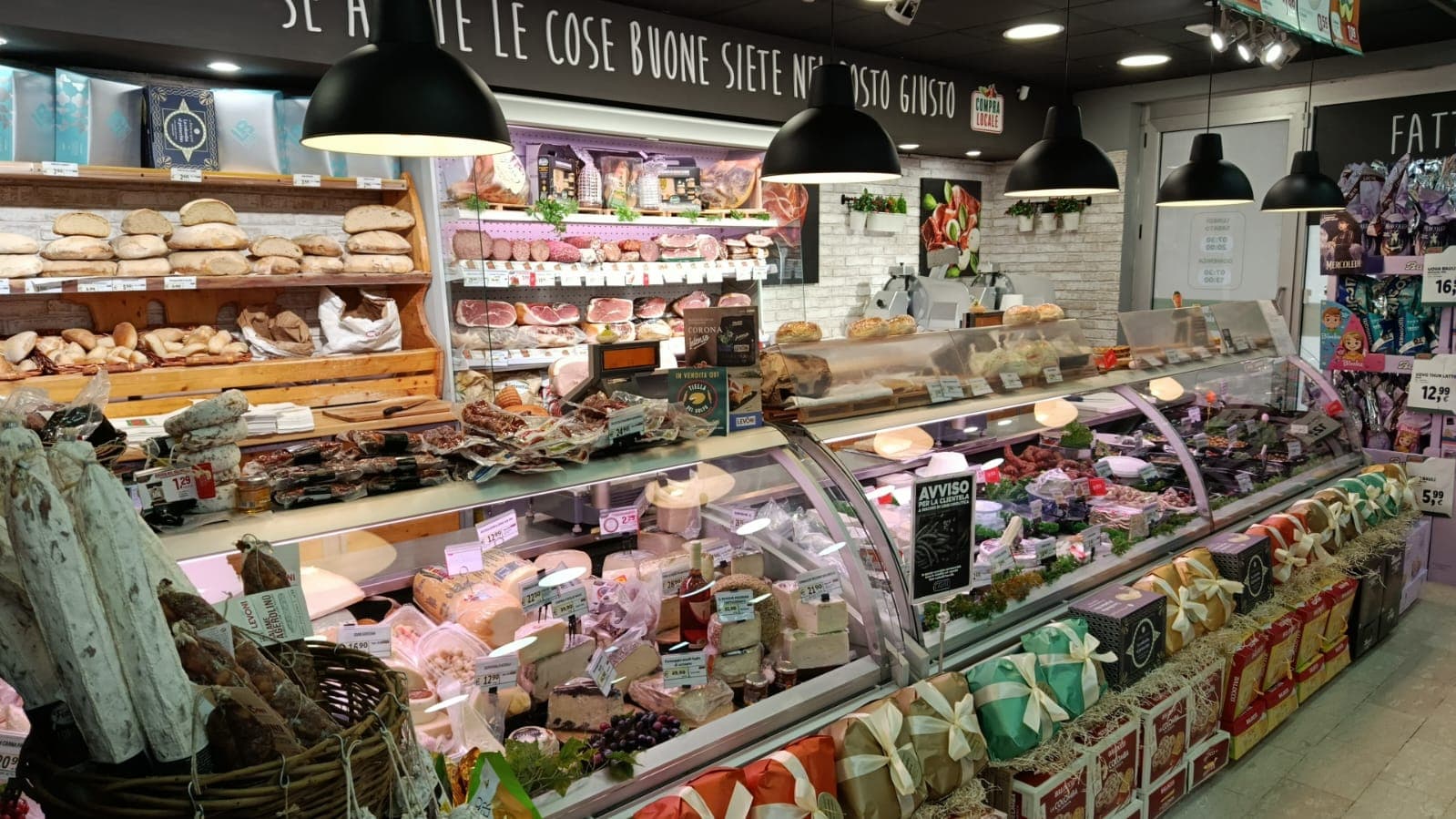 Bancone di una salumeria con pane fresco, formaggi e salumi esposti sotto moderne lampade nere.