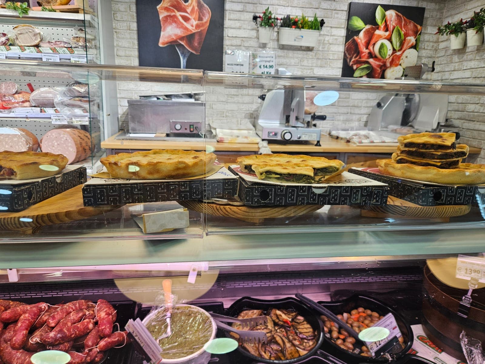 Bancone di una gastronomia con torte salate, salsicce, olive e verdure grigliate in esposizione.