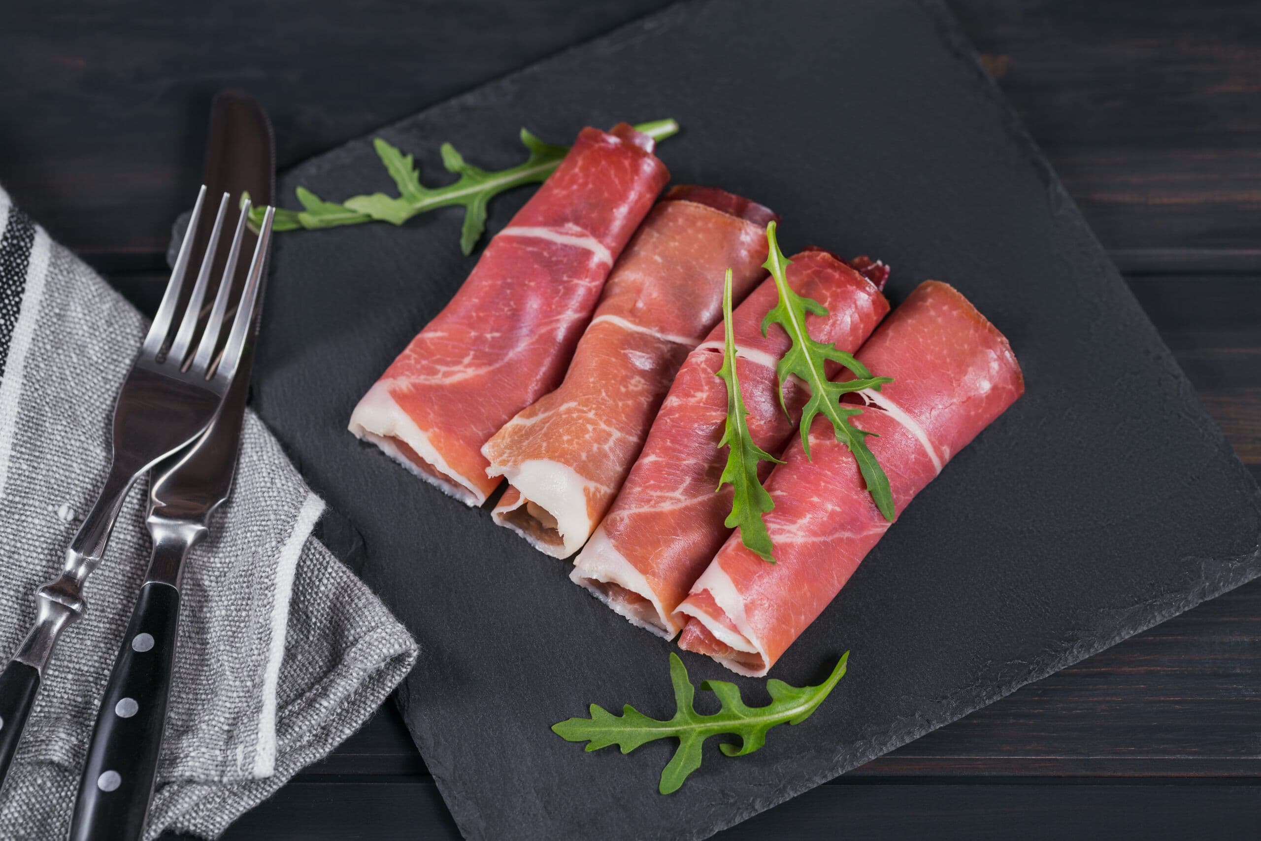Rotoli di prosciutto crudo su tagliere di ardesia nera con rucola fresca e posate.